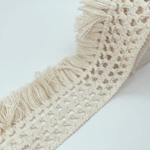 Op de afbeelding: Crèmekleurige macramé-bies met een franje rand. De bies is opgerold en toont het ingewikkelde geweven patroon en de zachte textuur van het katoenen materiaal. Deze decoratieve bies is geschikt voor diverse knutselprojecten.