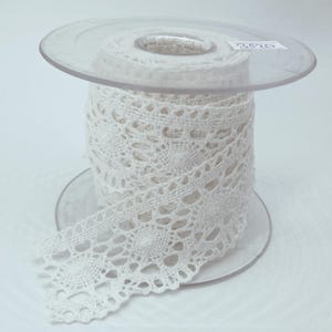 May include: A spool of white crochet lace trim featuring a repeating circular floral pattern. The lace is wound around a clear plastic bobbin with the number 3520 printed on a small white label attached to the top edge of the spool.