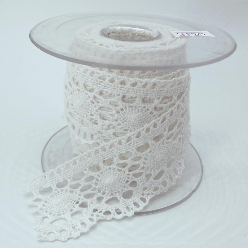 May include: A spool of white crochet lace trim featuring a repeating circular floral pattern. The lace is wound around a clear plastic bobbin with the number 3520 printed on a small white label attached to the top edge of the spool.