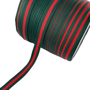 Op de afbeelding: Een rol groen lint met twee rode strepen in het midden. Het lint is strak opgerold, met een deel afgewikkeld. Dit lint is geschikt voor knutselen, cadeauverpakking of decoratieve doeleinden.
