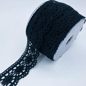 May include: A roll of black crochet lace trim featuring a repeating circular floral pattern with intricate openwork details. The lace is wound around a clear plastic spool, showcasing the continuous length of the decorative fabric trim.