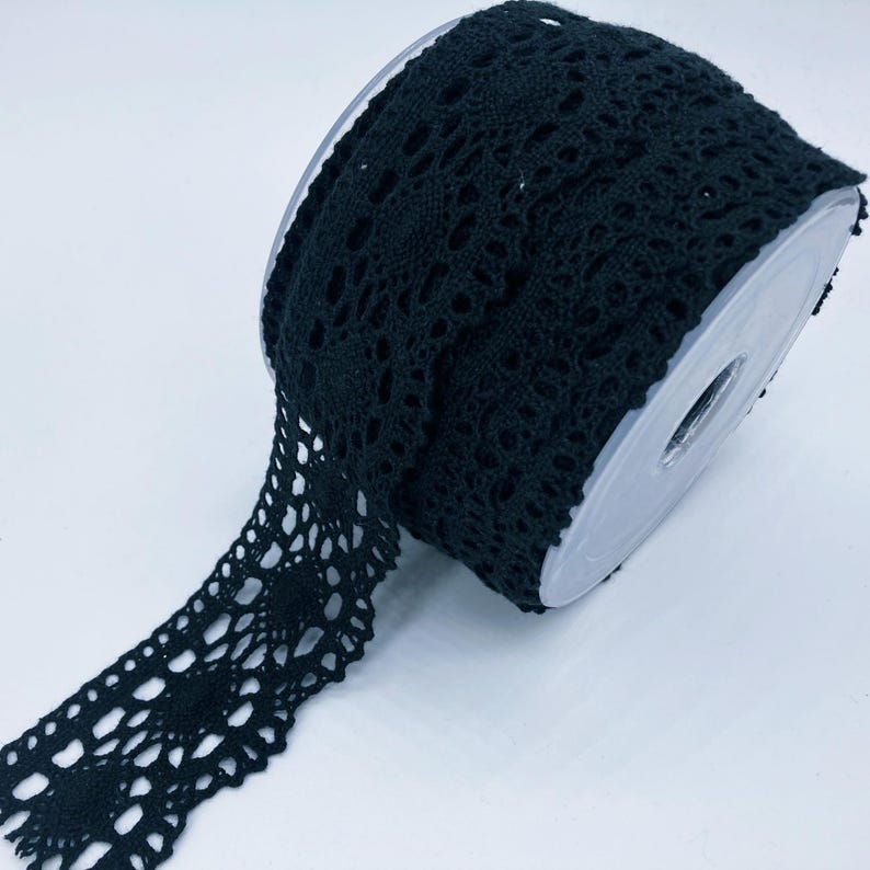 May include: A roll of black crochet lace trim featuring a repeating circular floral pattern with intricate openwork details. The lace is wound around a clear plastic spool, showcasing the continuous length of the decorative fabric trim.