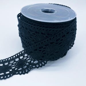 May include: A spool of black lace trim featuring a repeating circular floral pattern with scalloped edges. The lace is wound tightly around a clear plastic cylindrical core, with one end of the trim extending outward from the main roll.