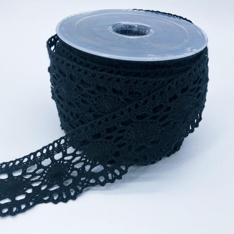 May include: A spool of black lace trim featuring a repeating circular floral pattern with scalloped edges. The lace is wound tightly around a clear plastic cylindrical core, with one end of the trim extending outward from the main roll.