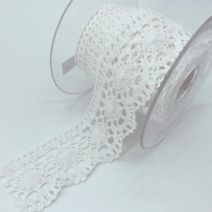 May include: A spool of white lace trim featuring a repeating circular floral pattern with scalloped edges. The lace is wound around a clear plastic spool, with a portion of the intricate, openwork fabric extending outward from the centre.
