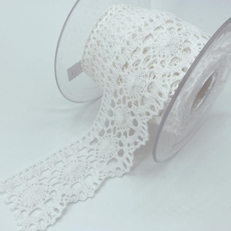 May include: A spool of white lace trim featuring a repeating circular floral pattern with scalloped edges. The lace is wound around a clear plastic spool, with a portion of the intricate, openwork fabric extending outward from the centre.