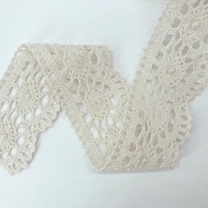 May include: A strip of off-white crochet lace trim featuring a repeating circular pattern with openwork detailing and scalloped edges. The lace has a textured, woven appearance with a consistent design throughout the length of the fabric trim.