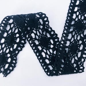 May include: A black crochet lace trim featuring a repeating floral pattern with intricate openwork details. The fabric strip displays a scalloped edge design and a continuous, interconnected mesh structure throughout the entire length of the material.