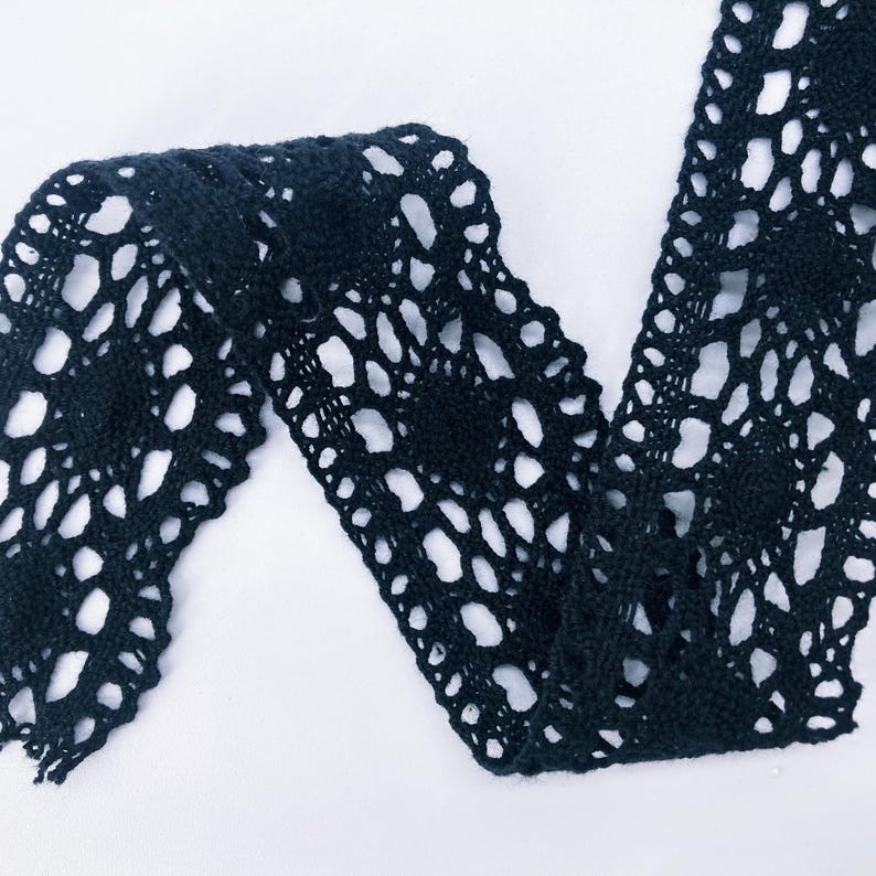 May include: A black crochet lace trim featuring a repeating floral pattern with intricate openwork details. The fabric strip displays a scalloped edge design and a continuous, interconnected mesh structure throughout the entire length of the material.