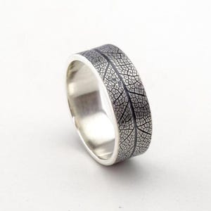 Könnte beinhalten: Ein silberner Ring mit einem Blattader-Muster. Der breite Ring ist mit einem detaillierten, dunkelgrauen Blattmuster verziert. Die metallische Oberfläche des Rings ist vor einem weißen Hintergrund platziert, was seine komplizierte Textur und sein poliertes Finish hervorhebt.