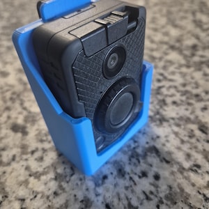 Puede incluir: Un soporte de plástico azul para una cámara corporal negra con una lente y un botón. La cámara tiene una superficie negra texturizada.