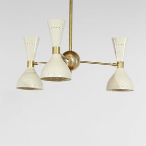 Op de afbeelding: Een messing en witte kroonluchter met vier lichten. De kroonluchter heeft een centrale messing bol en drie armen die naar de lichten uitstrekken. Elk licht heeft een witte, klokvormige kap met een messing bovenkant.