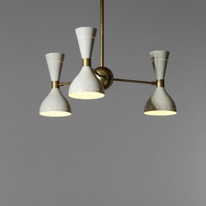 Op de afbeelding: Een messing en witte kroonluchter met vier lampen. De kroonluchter heeft een modern ontwerp met een centrale steel en drie armen die naar buiten uitstrekken. Elke arm heeft een witte, klokvormige kap met een kleine, ronde opening aan de bovenkant.