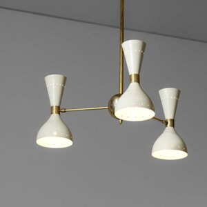 Op de afbeelding: Een messing en witte kroonluchter met vier lampen. De kroonluchter heeft een modern ontwerp met een centrale steel en drie armen die naar de zijkanten uitstrekken. Elke arm heeft een witte, klokvormige kap met een klein gaatjespatroon.