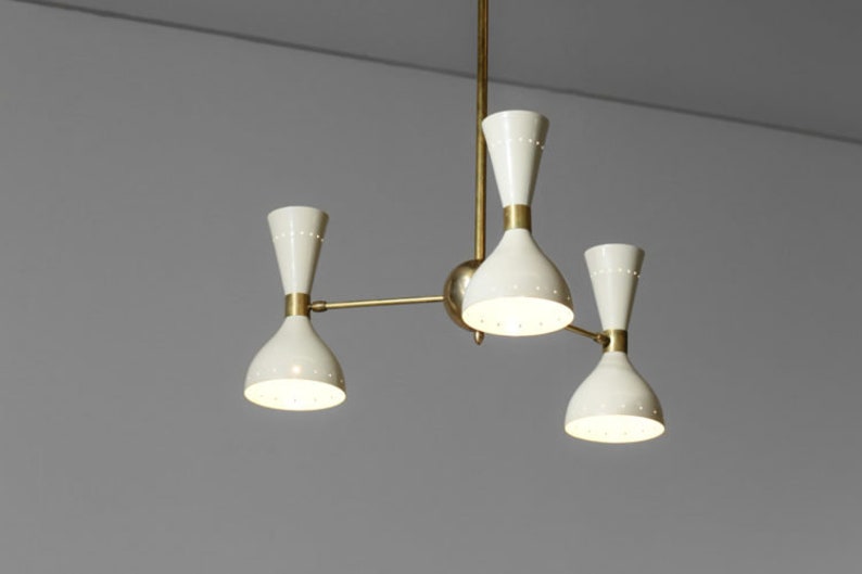 Op de afbeelding: Een messing en witte kroonluchter met vier lampen. De kroonluchter heeft een modern ontwerp met een centrale steel en drie armen die naar de zijkanten uitstrekken. Elke arm heeft een witte, klokvormige kap met een klein gaatjespatroon.