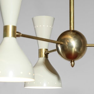 Op de afbeelding: Een messing en witte kroonluchter met vijf lampen. De kroonluchter heeft een centrale bol en vier armen die naar de lampen uitstrekken. De lampen zijn wit en hebben een kegelvorm met een klein gaatje bovenaan.