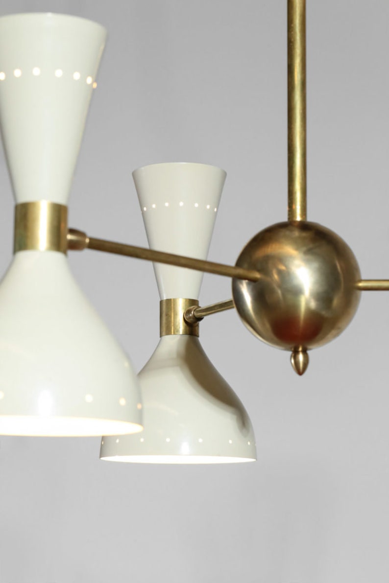 Op de afbeelding: Een messing en witte kroonluchter met vijf lampen. De kroonluchter heeft een centrale bol en vier armen die naar de lampen uitstrekken. De lampen zijn wit en hebben een kegelvorm met een klein gaatje bovenaan.