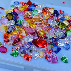 Puede incluir: Una colección de piedras preciosas facetadas sueltas en varias formas y tamaños, incluyendo cortes redondos, ovalados, de pera y cuadrados. Las piedras muestran un espectro vibrante de tonos rojos, azules, verdes, amarillos, morados y transparentes.