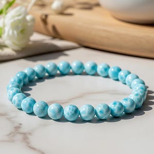 Op de afbeelding: Een lichtblauwe kralenarmband met een gemarmerd patroon. De armband is gemaakt van ronde kralen en wordt gepresenteerd op een wit en grijs marmeren oppervlak. Het is een sieraad.