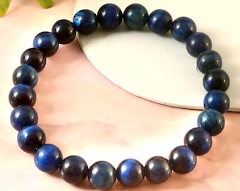 May include: A bracelet made of dark blue and black spherical beads. The beads are strung together to form a circular shape. The bracelet is displayed on a light pink surface with a white, curved edge.
