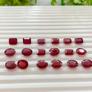 Puede incluir: Una variedad de gemas pulidas de color rojo intenso en varias formas, incluyendo cuadrados, rectángulos y óvalos. Las gemas están dispuestas sobre una superficie blanca y acanalada, con follaje verde en el fondo. Las gemas son probablemente rubíes.