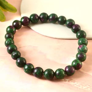 May include: A bracelet made of green and maroon gemstone beads. The beads are round and polished. The bracelet is displayed on a light pink surface with a white accent.