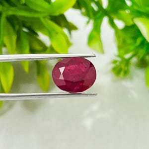 Pode incluir: Uma pedra preciosa de rubi facetada de formato oval com uma cor vermelha profunda, segurada por uma pinça de metal prateado. A gema exibe múltiplas superfícies cortadas que refletem a luz, destacando sua qualidade translúcida e tom vermelho vibrante.