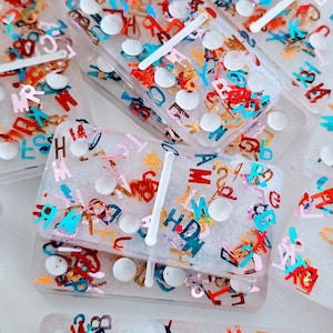 May include: A set of white dominoes with a clear, translucent finish. Each domino is covered in colorful confetti-like letters and numbers. The dominoes are stacked on top of each other.