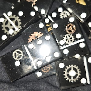May include: Black dominoes with white dots and decorative metal gears. The dominoes are scattered on a dark fabric surface.