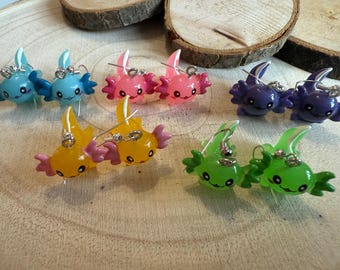 Peut inclure: Boucles d'oreilles axolotl colorées en bleu, rose, violet, jaune et vert. Chaque boucle d'oreille présente un design d'axolotl souriant avec des crochets d'oreilles argentés. Les boucles d'oreilles sont présentées sur une surface en bois.
