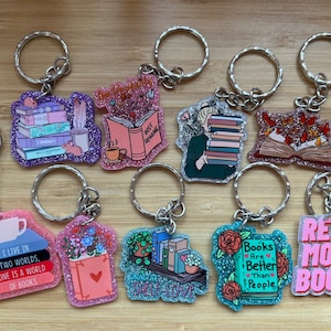 May include: Nine different book-themed keyrings with glitter accents. The keyrings feature various designs, including a stack of books, a book with a cup of coffee, a person holding a stack of books, a book with butterflies, a stack of books with a cup of tea, a book with flowers, a bookshelf with plants, a book with roses, and a book with the text "Read More Books".