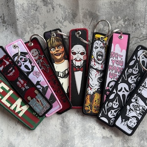 May include: A collection of rectangular fabric keychains featuring various horror movie themes, including characters like Billy the Puppet, Art the Clown, and Ghostface, with text such as ELM, COME PLAY WITH US, I WANT TO PLAY, and NO YOU HANG UP.