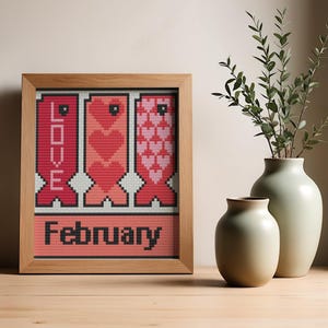 May include: A framed cross-stitch artwork with the word "LOVE" vertically and "February" horizontally. The artwork features red and pink hearts and is set in a wooden frame. Two vases with greenery are in the background.