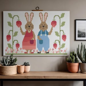 Puede incluir: Una obra de arte de punto de cruz con dos conejos sosteniendo fresas. Uno lleva un vestido rojo, el otro un peto azul. Enredaderas verdes con fresas rojas y flores enmarcan los conejos. La obra se exhibe sobre una mesa de madera.