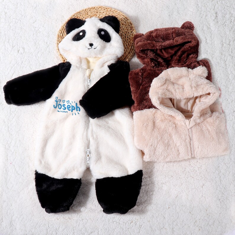May include: A white and black panda-themed baby jumpsuit with a hood, zipper, and the name "Joseph" in blue. Next to it are two hooded jackets, one brown and one beige, with bear-like ears. The items are made of soft, plush material.