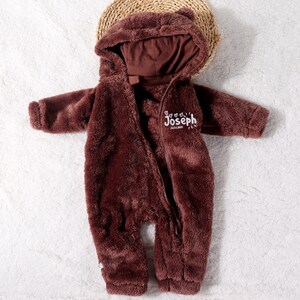 May include: A brown, plush baby onesie with a hood and a zipper closure. The onesie has a bear-like design with small ears on the hood. The name "Joseph" is embroidered on the chest. The onesie is lying on a white surface.