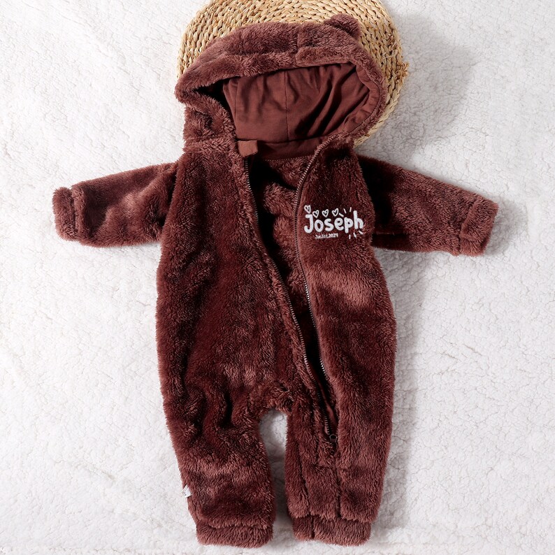 May include: A brown, plush baby onesie with a hood and a zipper closure. The onesie has a bear-like design with small ears on the hood. The name "Joseph" is embroidered on the chest. The onesie is lying on a white surface.