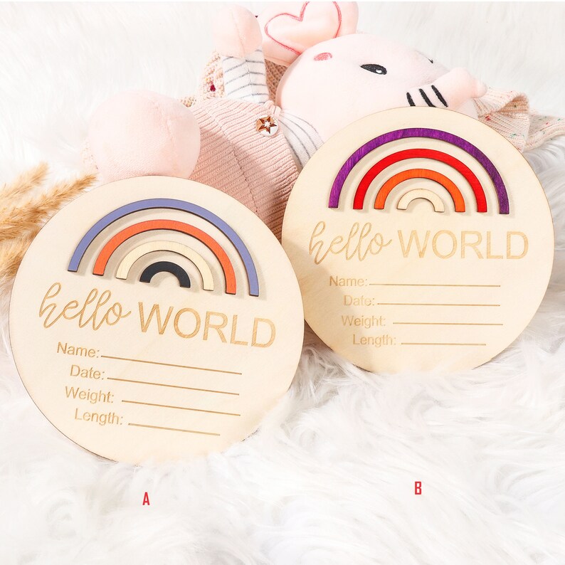 May include: Two round wooden baby announcement plaques. Each plaque has a rainbow design in shades of purple, orange, and blue. The words "hello WORLD" are engraved on each plaque, along with lines for Name, Date, Weight, and Length.