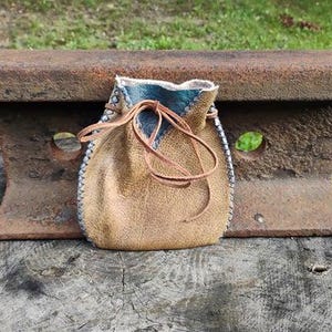 May include: A handcrafted leather pouch with a drawstring closure. The pouch is tan with a blue interior and white stitching along the edges. The drawstring is a lighter brown. The pouch is sitting on a rustic metal object.