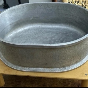 May include: An oval-shaped silver-colored metal roasting pan with a textured exterior finish and smooth interior. The pan features two integrated handles on opposite sides, each with a distinct notched design for gripping.