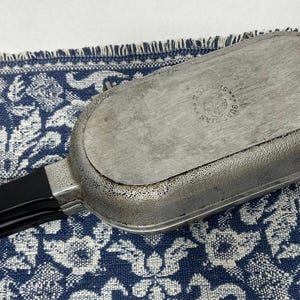 May include: An oval-shaped metal pan with a textured silver finish and a black handle. The base of the pan features a circular stamp with the text Guardian Service. The pan is constructed from two joined metal sections with a visible seam.