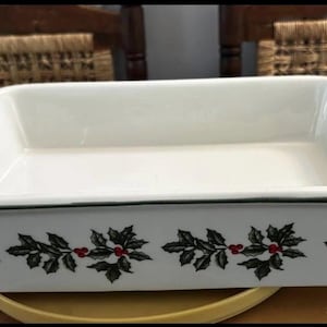 May include: A rectangular white ceramic baking dish featuring a repeating pattern of green holly leaves and small red berries around the exterior side walls. The dish has integrated handles on both ends and a smooth, glossy interior finish.