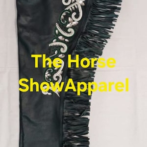 May include: Green leather chaps with fringe and silver and green embroidery. The chaps have a strap with a buckle and a metal button. The text "The Horse Show Apparel" is visible on the chaps.