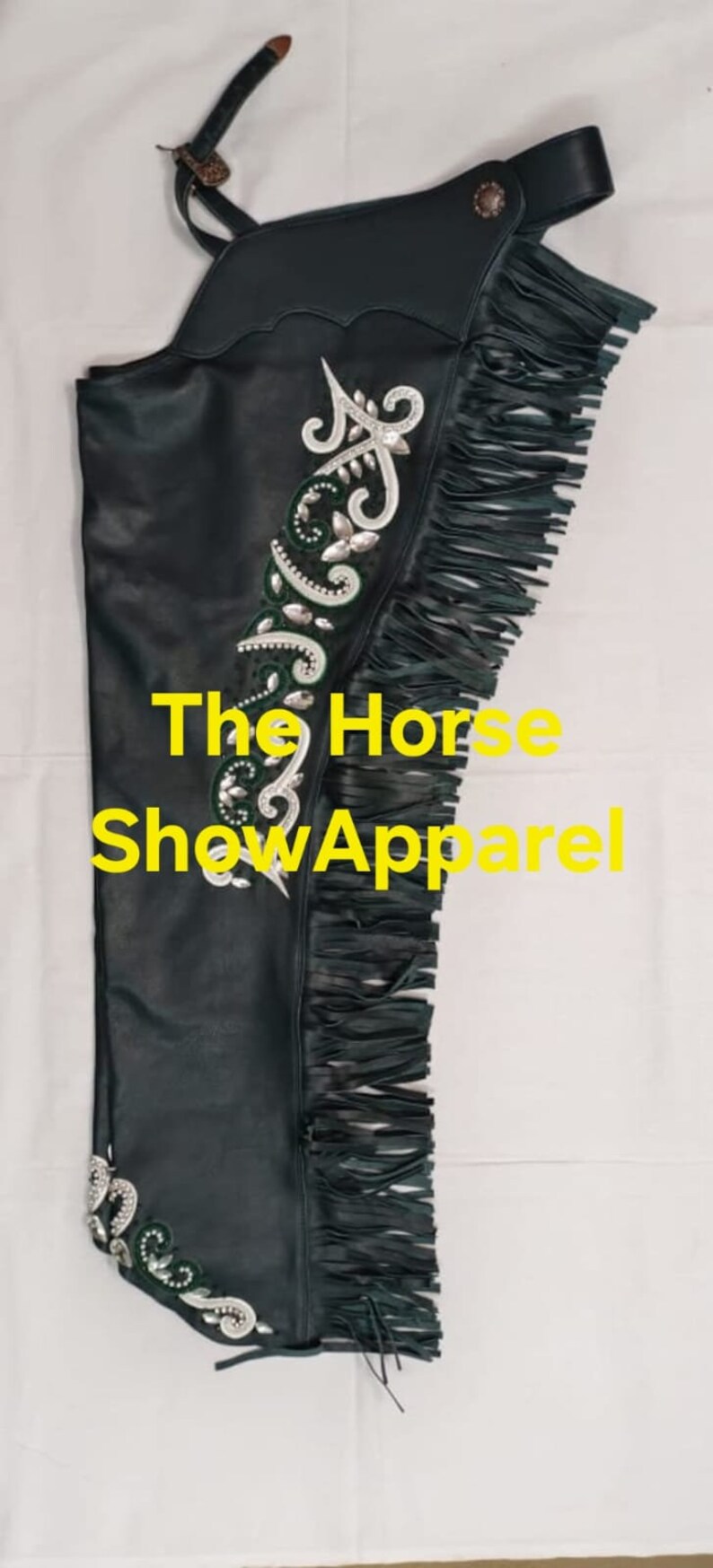 May include: Green leather chaps with fringe and silver and green embroidery. The chaps have a strap with a buckle and a metal button. The text "The Horse Show Apparel" is visible on the chaps.