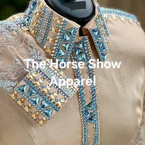 May include: A beige button-up shirt with a blue and gold rhinestone design on the collar and cuffs. The shirt has a lace overlay on the sleeves. The text "The Horse Show Apparel" is visible on the shirt.