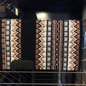 Könnte beinhalten: Schwarz-weißer Sattelpad mit orangefarbenen und kupferfarbenen Akzenten. Der Sattelpad hat ein geometrisches Muster und hängt an einem Metallgestell.