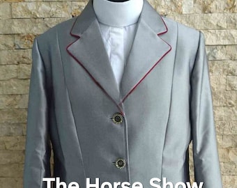 May include: A silver riding jacket with a red trim. The jacket has a single breasted closure with three buttons. The jacket has a pocket on the left side. The text "The Horse Show Apparel" is printed on the jacket.