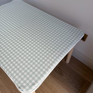 May include: A small, rectangular table with a light green and white gingham pattern top. The table has a white trim and four light-colored wooden legs. The table is approximately 30 inches long and 20 inches wide.