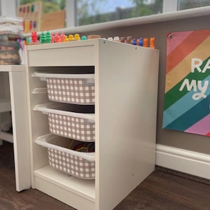 May include: A white storage unit with three pull-out bins featuring a brown and white checkered pattern. The top of the unit displays colorful toys. A rainbow-themed poster is visible in the background.