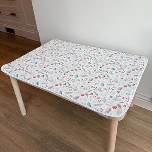 Può includere: Un tavolino rettangolare con una superficie bianca decorata con un motivo botanico di foglie, bacche e funghi in colori tenui. Il tavolo ha angoli arrotondati e gambe in legno chiaro. Il tavolo è lungo circa 61 cm.